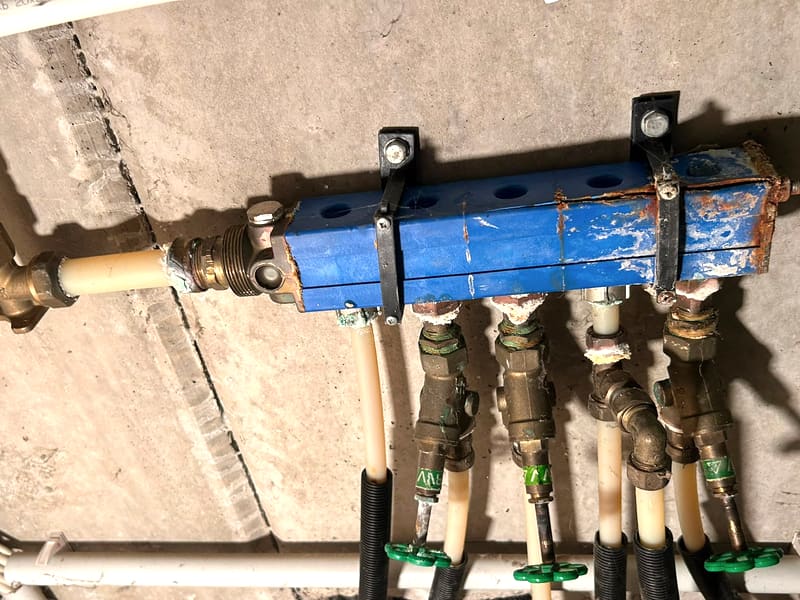 Alter Wasserverteiler aus Stahl an einer Betonwand, an den Enden durchgerostet, an den Verschraubungen weiße Kalkablagerungen.